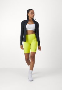 Jachetă neagră cu fermoar, sutien sport alb și pantaloni scurți ciclist galbeni strălucitori cu fermoare pe lateral. Modelul poartă adidași albi și șosete până la glezne.