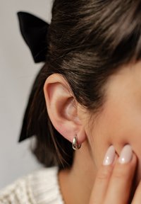 Srebrna kolczyk w kształcie obręczy przymocowany do przekłutego ucha, charakteryzujący się gładkim, zaokrąglonym wzorem. Ucho ma proste ciemne włosy i czarną kokardkę.