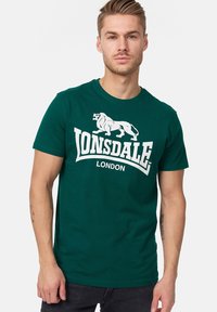 Zöld pamut póló, fehér oroszlán grafikával és a "LONSDALE LONDON" felirattal a mellkasán. Rövid ujjakkal és kerek nyakkivágással.