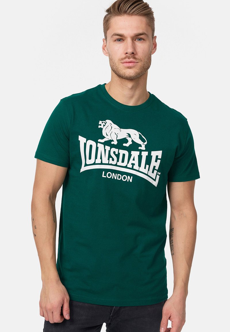 Zöld pamut póló, fehér oroszlán grafikával és a "LONSDALE LONDON" felirattal a mellkasán. Rövid ujjakkal és kerek nyakkivágással.