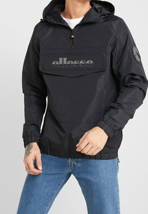 Mann trägt schwarzen ellesse Kapuzen-Windbreaker mit Fronttasche und blauer Jeans, Handtattoo am linken Handgelenk sichtbar.