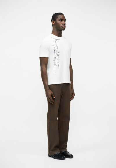 Άνδρας που στέκεται φορώντας λευκό πουκάμισο με κοντά μανίκια και κάθετη μαύρη γραφή, καφέ παντελόνι φαρδιάς γραμμής και μαύρα παπούτσια.