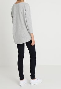 Top gris de manga larga con dobladillo redondeado, combinado con jeans ajustados oscuros y zapatillas blancas, visto desde atrás. Textura de tela suave.
