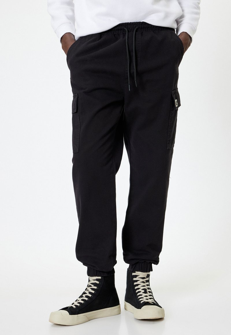 Sorte cargo-bukser med elastisk talje, sidelommer og opsmøgede afslutninger. Båret med højtstående sorte sneakers og en hvid sweatshirt.