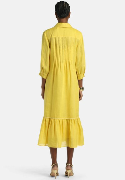 Žena s pletenými vlasmi má na sebe žlté midi šaty s dlhým rukávom s záhybmi na zadnej strane a riaseným lemom, doplnené béžovými opätkami.