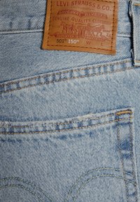 Ανοιχτό μπλε τζιν παντελόνι με κλασικό σχέδιο, που διαθέτει καφέ δερμάτινη ετικέτα και κίτρινη ραφή στην πίσω τσέπη. Ονομασία μοντέλου 501.