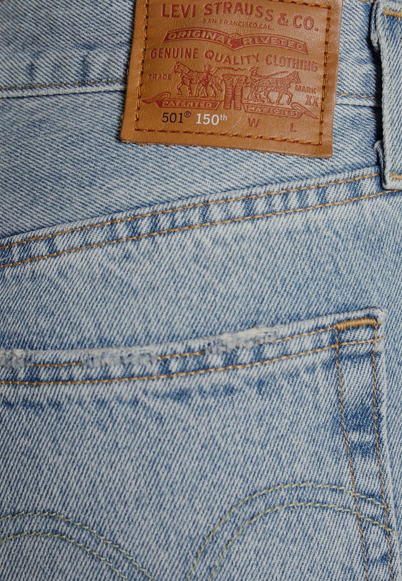 Jeans de denim azul claro con un diseño clásico, que presentan una etiqueta de cuero marrón y costuras amarillas en el bolsillo trasero. Designación del modelo 501.