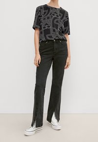 Person iført sort grafisk top, mørkegrå jeans med benåbninger foran, og hvide platform sneakers, stående mod en ensfarvet lys baggrund.