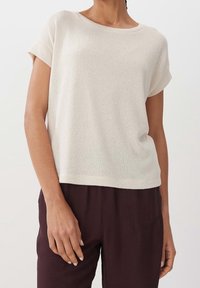 Ljusbeige stickad kortärmad topp med en bred rund halsringning och en avslappnad passform, tillsammans med mörkbruna byxor. Mjuk textur och minimalistisk design.