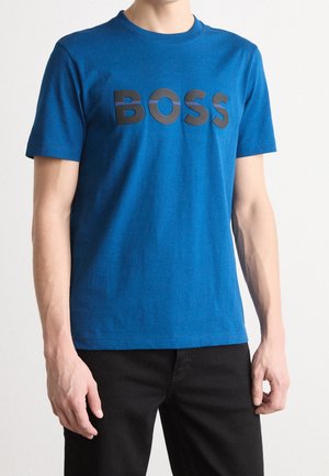 Man draagt een blauw T-shirt met korte mouwen met zwarte hoofdletters "BOSS" over de borst, gecombineerd met een zwarte broek.