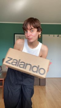 Kartonowe pudełko Zalando trzymane w rękach, prostokątny kształt, jasnobrązowy kolor z czarnym logo. W tle widać zieloną ścianę i meble.