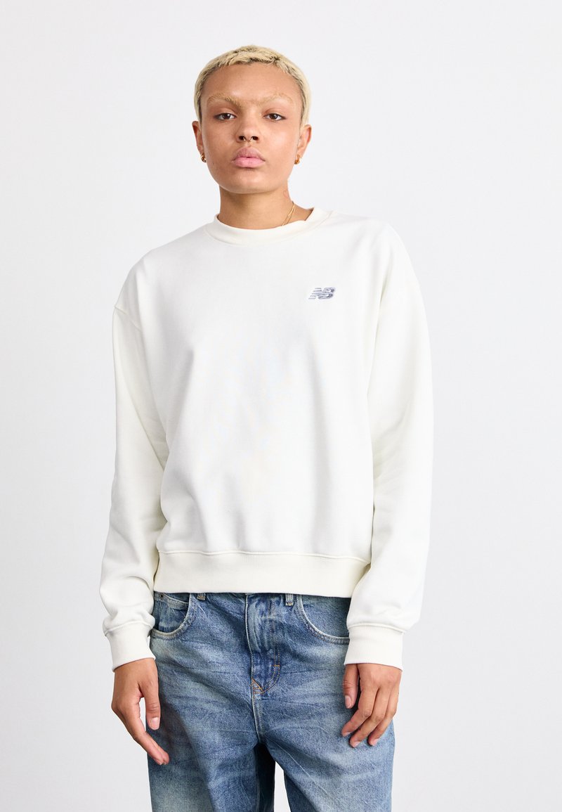 Sudadera blanca de cuello redondo con puños y dobladillo acanalados, que presenta un pequeño logo gris en el pecho. Llevada con jeans azules claros y desgastados.
