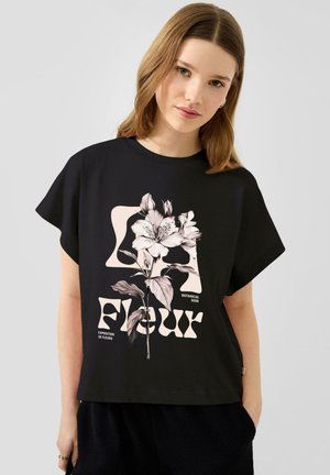 Jonge vrouw die een zwart T-shirt draagt met een grote beige bloemenprint en de tekst "Fleur" in gestileerd lettertype op de voorkant.