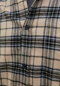 Karierte Flanellbluse in Tan mit Schwarz- und Graufarbigen Checkmustern. Verfügt über einen V-Ausschnitt und schwarze Knopfdetails auf der Brust. Weiche Textur.
