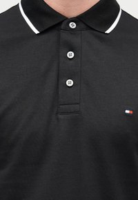 Μαύρο βαμβακερό πόλο με κλασικό γιακά, λευκή ρίγα, πατιλέτα με τρία κουμπιά και μικρό κεντημένο λογότυπο σε κόκκινο, άσπρο και μπλε.