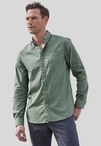 Camisa verde de botones hecha de algodón, con un bolsillo en el pecho, mangas largas y un corte ligeramente relajado. Combinada con pantalones oscuros.