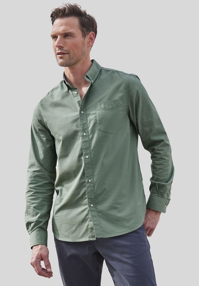 Camisa verde de botones hecha de algodón, con un bolsillo en el pecho, mangas largas y un corte ligeramente relajado. Combinada con pantalones oscuros.