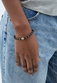 Bracciale di perle nere con accento di croce dorata, indossato su una mano, sopra dei jeans di denim. Le perle variano in dimensione, creando un aspetto testurizzato.