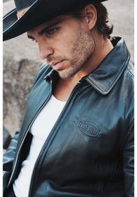 Veste en cuir noir avec fermeture éclair, dotée d'un col classique et d'un logo en relief. Portée sur un débardeur blanc à la finition texturée.