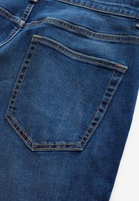 Achterzak van blauwe denim jeans met zichtbare oranje stiksels en stoftextuur tegen een witte achtergrond.