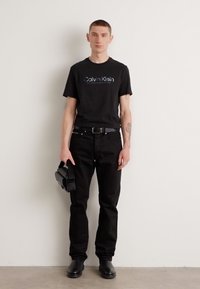 Sort bomulds t-shirt med "Calvin Klein" logo, parret med sorte denim jeans, sort bælte og en lille sort taske. Glat tekstur og tætsiddende silhuet.