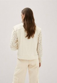 Beiger Denim-Weste mit breiten Schultern und geradem Schnitt, über einer gemusterten hellen Bluse mit Blumenmotiven und Puffärmeln getragen.