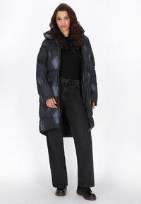 Geplüsterter, dunkelgrauer langer Mantel mit Kapuze, der eine glänzende, gestreifte Textur und einen Frontreißverschluss aufweist; das Modell trägt ein schwarzes Oberteil, Jeans und Stiefel.