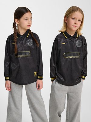 Zwei Kinder in schwarzen Sporttrikots mit langen Ärmeln und grauen Jogginghosen stehen vor einem schlichten weißen Hintergrund.