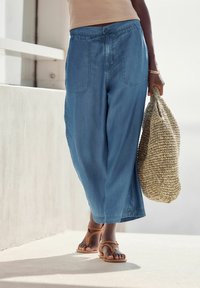 Pantaloni relaxați de culoare albastru deschis, cu două buzunare frontale, purtați cu sandale maro cu fâșii și ținând o geantă țesută de culoare bej.
