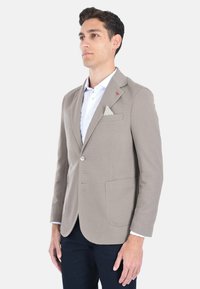 Blazer beige in tessuto testurizzato, chiusura a singolo bottone e rever a punta. Caratterizzato da un fazzoletto bianco nella tasca e un sottile accento rosso sul rever.
