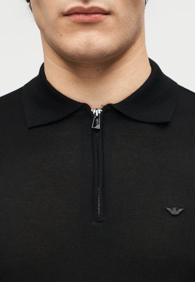 Κοντινό πλάνο ενός ατόμου που φοράει μαύρο πουκάμισο με φερμουάρ και γιακά, με ένα μικρό κεντητό λογότυπο στο αριστερό μέρος του στήθους.