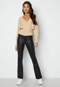 Beige wikkeltrui met brede mouwen en een V-hals, gecombineerd met strakke zwarte neplederen flared broeken en witte sneakers.