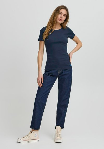 Tricou ajustat de culoare albastru marin, cu mâneci scurte, purtat cu blugi închisi la culoare și adidași din pânză bej cu logo-ul în formă de stea pe lateral.