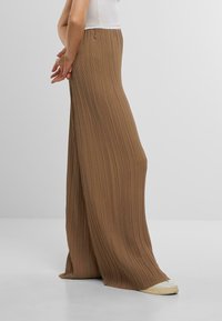 Brede, geplooide broek in een rijke, bruine stof. Kenmerkt zich door verticale pinch-pleats en een fitted tailleband, gecombineerd met een witte schoen.