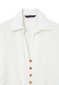 Camicia bianca con colletto in materiale morbido, dotata di una chiusura a bottoni sul davanti con cinque bottoni rotondi marroni e un tessuto testurizzato.