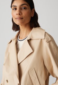 Trench coat beige con un ampio colletto, chiusura doppiopetto e due tasche laterali. Realizzato in tessuto liscio con una leggera lucentezza.