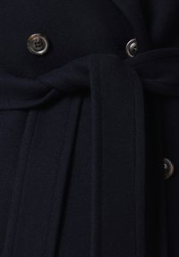 Granatowy płaszcz wełniany z paskiem w talii, podwójnym zapięciem na guziki i okrągłymi dekoracyjnymi detalami przypominającymi guziki, o subtelnym połysku.