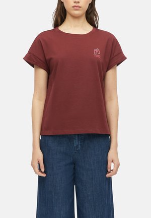 Frau trägt ein dunkelrotes T-Shirt mit hochgekrempelten Ärmeln und kleinem Flip-Flop-Motiv über dem Text auf der linken Brust, kombiniert mit weitem blauen Jeans.