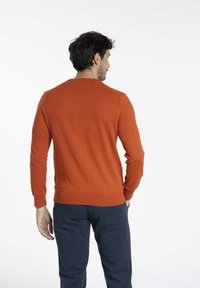 Pull en tricot orange avec un col rond, des poignets et un ourlet côtelés, vu de dos, associé à un pantalon sombre sur fond blanc.