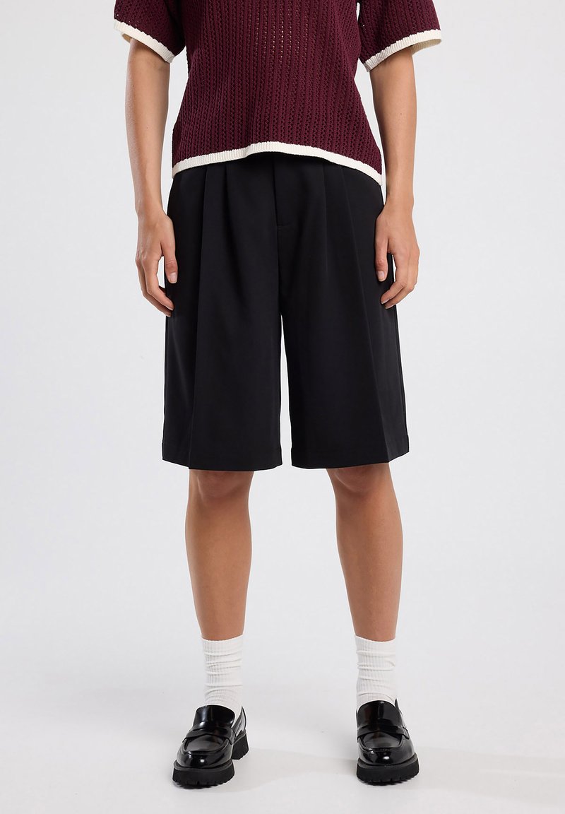Black tailored shorts with pleats, worn with a burgundy textured knit top, featuring white trim. Styled with white socks and black loafers.