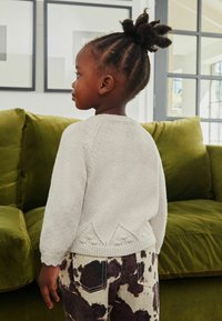 Lys grå strikket sweater med bølget kantdetalje, parret med brune og hvide mønstrede bukser, mod en baggrund af en grøn fløjlssofa.