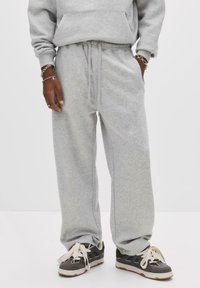Person iført grå sweatpants og matchende h�ættetrøje med sorte sneakers, der har hvide snørebånd, stående med hænderne i lommerne.