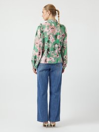 Bloemige blouse in groen, met roze en zwarte bloemen, lange mouwen en een iets losse pasvorm, gecombineerd met blauwe wijde jeans en hakken.