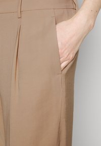 Mano dentro del bolsillo delantero de pantalones tailored beige con trabillas y un pliegue frontal.