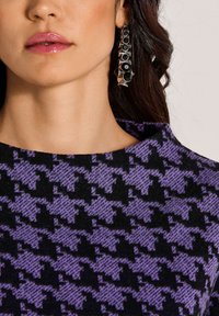 Vrouw met grote zilveren oorbellen met zwarte stenen en een paars-zwart hounds-tooth patroon top, met glanzend roze lippen.
