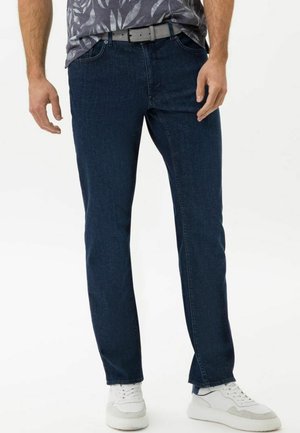 Jeans de mezclilla azul oscuro con corte recto, que cuentan con bolsillos frontales y un cinturón, combinados con zapatillas blancas. La textura parece lisa.