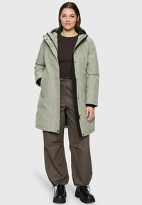 Femme portant un parka à capuche gris clair, un haut à manches longues noir, un pantalon à cordon brun et des bottes noires, se tenant devant un fond uni.