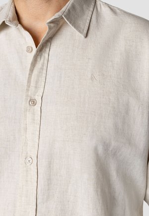 Jasnobeżowa lniana koszula z kołnierzykiem na guziki, plisą z przodu i subtelnym haftowanym akcentem na lewej części klatki piersiowej.