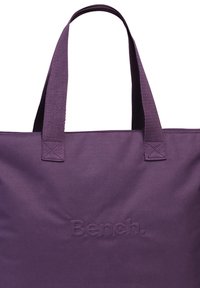 Paarse tote bag met twee stoffen handvatten, gemaakt van tekstielmateriaal en met een reliëf 'Bench.' logo aan de voorkant. Eenvoudig rechthoekig ontwerp.