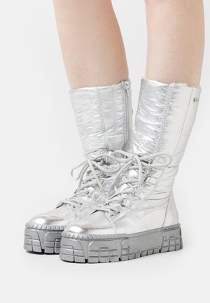Botas plateadas metálicas con textura acolchada, caña alta y diseño de cordones. Suelas de goma gris gruesas con detalles estriados. Cierre lateral con cremallera.